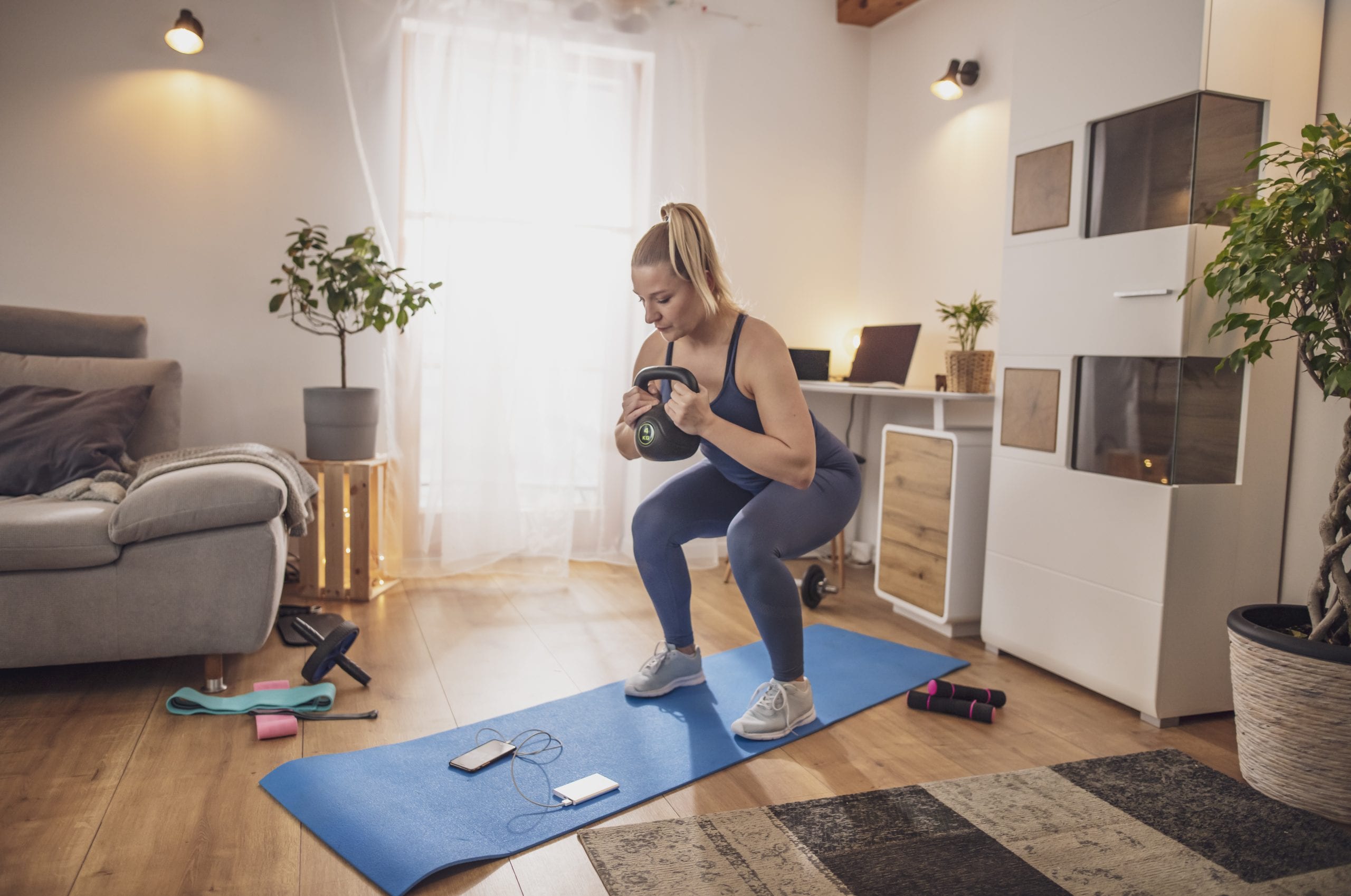 Young woman watching smart phone and squatting with kettlebell on yoga mat in living room