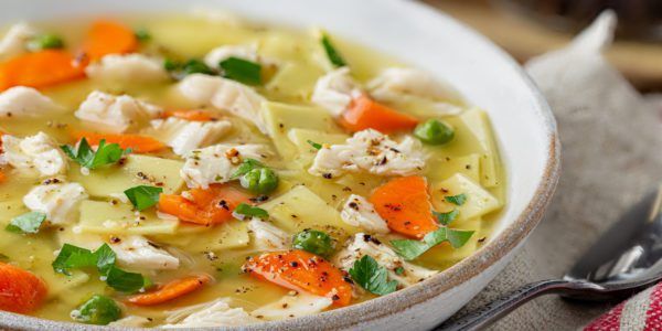 A white bowl full of chicken noodle soup with greens and carrots.