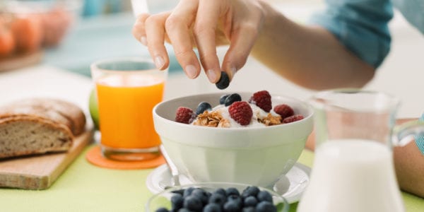 Woman having an healthy delicious breakfast at home with yogurt, cereals and fresh fruit, she is picking a blueberry