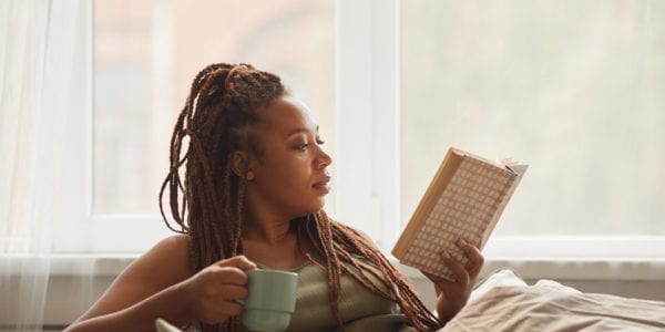 African young woman lying on sofa drinking coffee and reading an interesting book at home