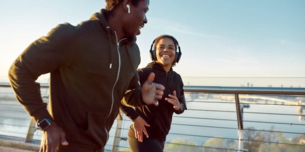 Morning is the best time for jogging. Young happy african couple or friends wearing headphones running together on the bridge, they talking and smiling during the run