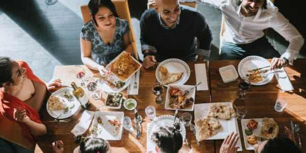 A group of adult friends and family of Indian ethnicity sit a table with a spread of delicious traditional food. A high angle view from overhead of the food, plates, and dishes at the dining table. Shot in Bellevue, Washington, United States.