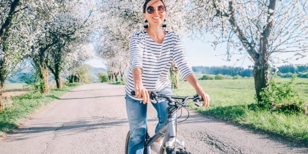 Happy, young woman rides her bike down a dirt road surrounded by blossoming trees and green fields.