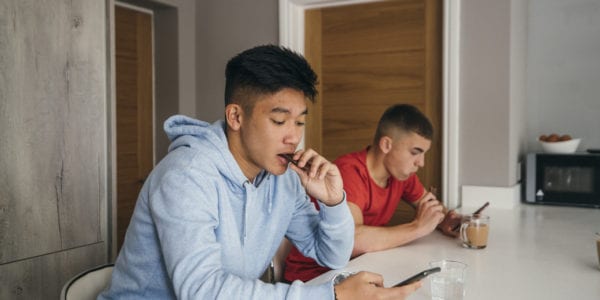 Two teenage boys sitting at a kitchen island while eating snacks. They are both looking at their phones while using social media.