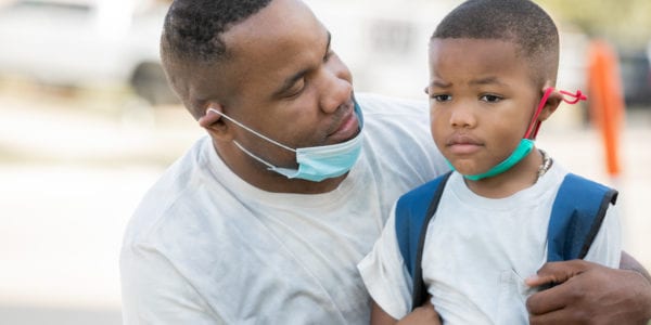 Father consoles anxious kindergartener while they wear masks as he's dropping him off for first day of school,