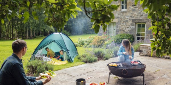 A shot of a young man and woman preparing a barbecue on a patio in the garden and their two young boys, one of which has downs syndrome are playing in a tent on the grass.