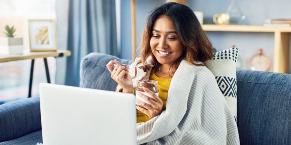 Shot of a woman looking at something on her laptop while eating chocolate from a jar