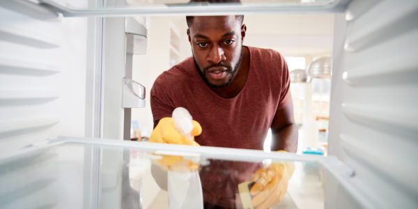 View Looking Out From Inside Empty Refrigerator As Man Wearing Rubber Gloves Cleans Shelves