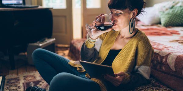 A woman drinks a glass of wine while looking at a smart device.