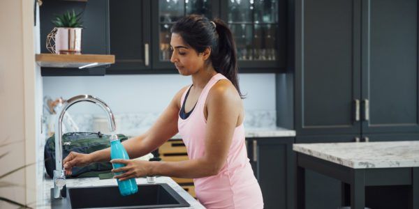 A woman stands at a kitchen sink filling her teal water bottle.