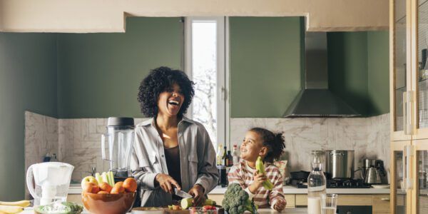 An African-American Single Mother Preparing Vegan Lunch In The Kitchen And Smiling With Her Little Daughter